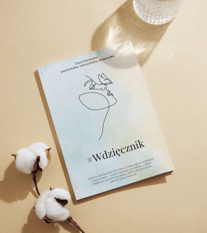 Książka zatytułowana „#Wdzięcznik” z abstrakcyjną ilustracją twarzy na okładce leży na beżowej powierzchni obok gałązek bawełny i szklanki wody.